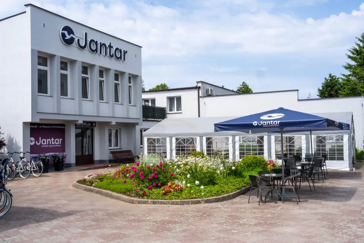 Ośrodek wypoczynkowy Jantar zaprasza do swojego ogrodu z kwiatami i stolikami pod parasolem. Obok recepcji stoją rowery.