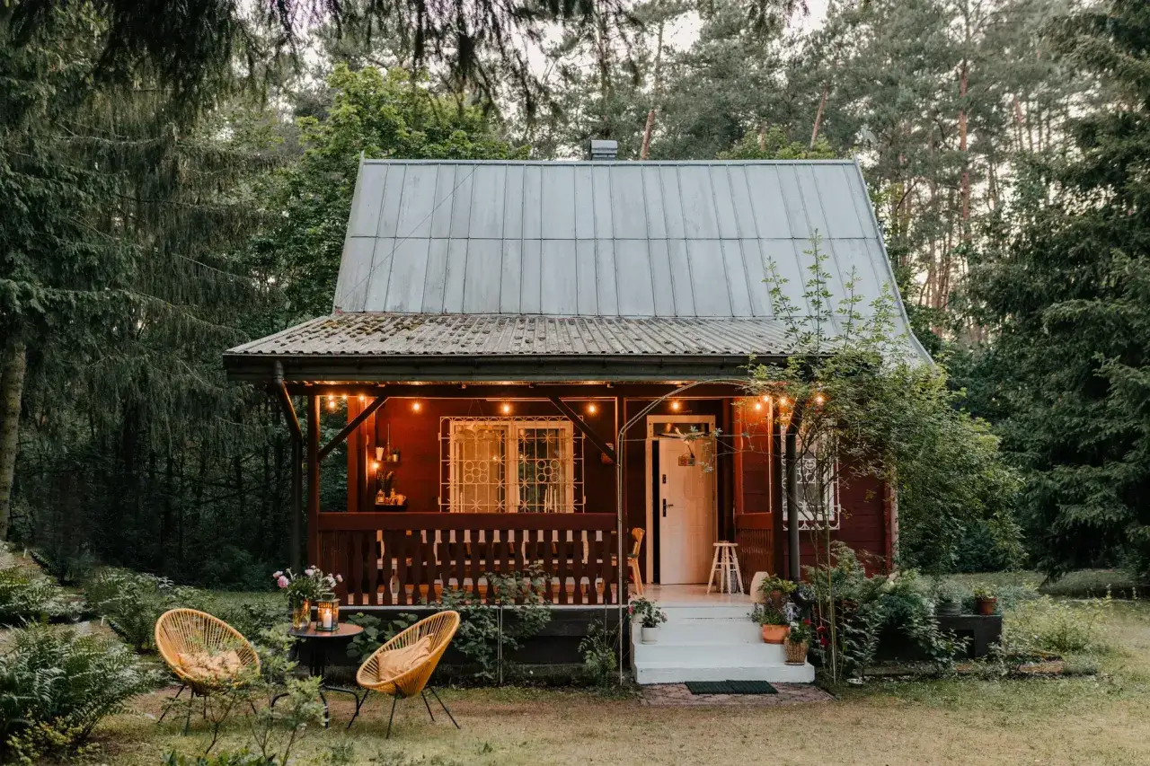 Urokliwy domek w lesie, z werandą oświetloną girlandami. Dwa żółte fotele zapraszają do odpoczynku. Idealne miejsce na relaks co oprócz booking.