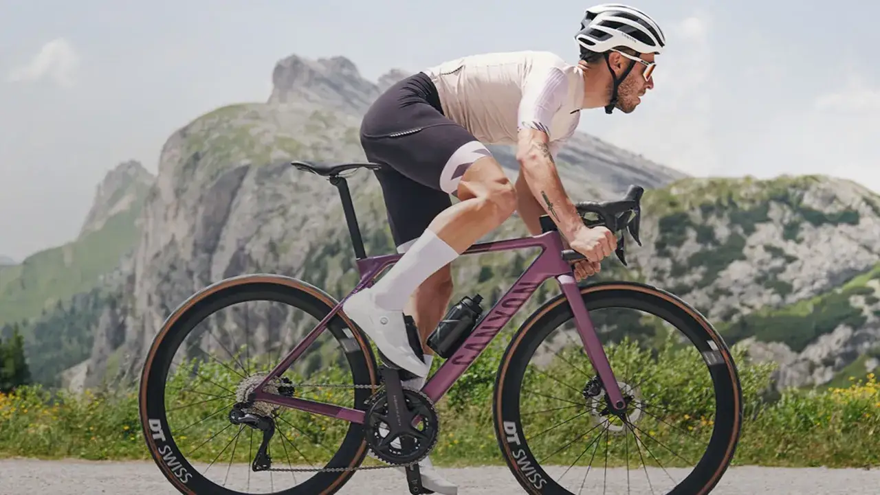 Kolarz w kasku i okularach ściga się na fioletowym rowerze szosowym po górskiej drodze. Tor rowerowy w tle.