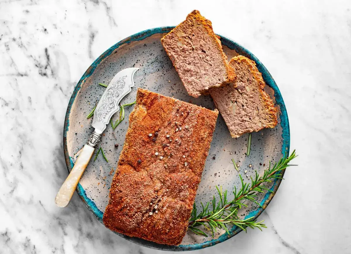 Pyszny pasztet Ani gotuje się na talerzu z gałązką rozmarynu i ozdobnym nożem. Dwa kawałki już pokrojone, kuszą apetycznym wyglądem.