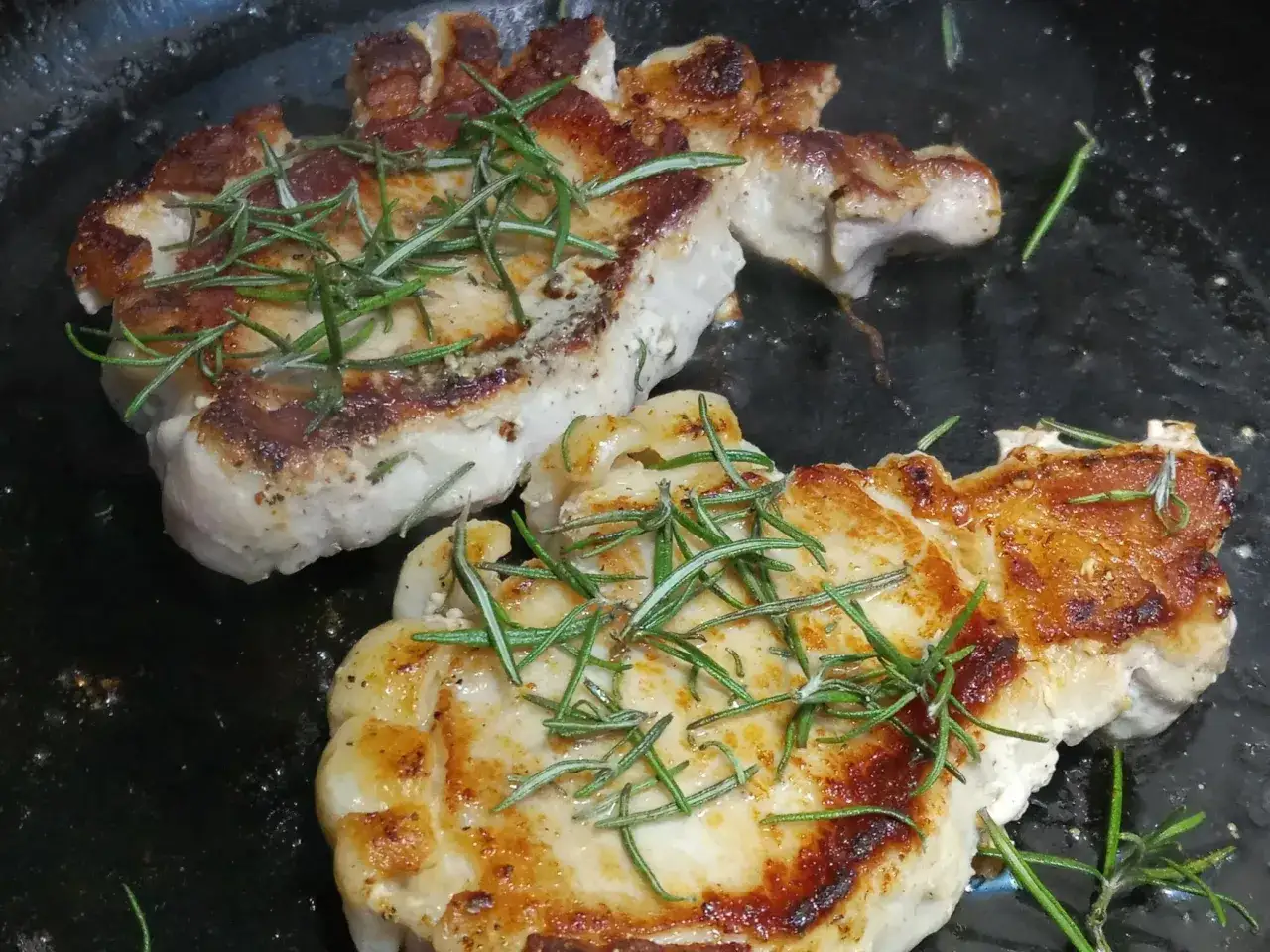 Dwa soczyste schabowe w trakcie smażenia na patelni, ozdobione gałązkami rozmarynu. Sekret jak usmażyć schabowego żeby panierka nie odpadała na patelni tkwi w odpowiedniej temperaturze i technice.