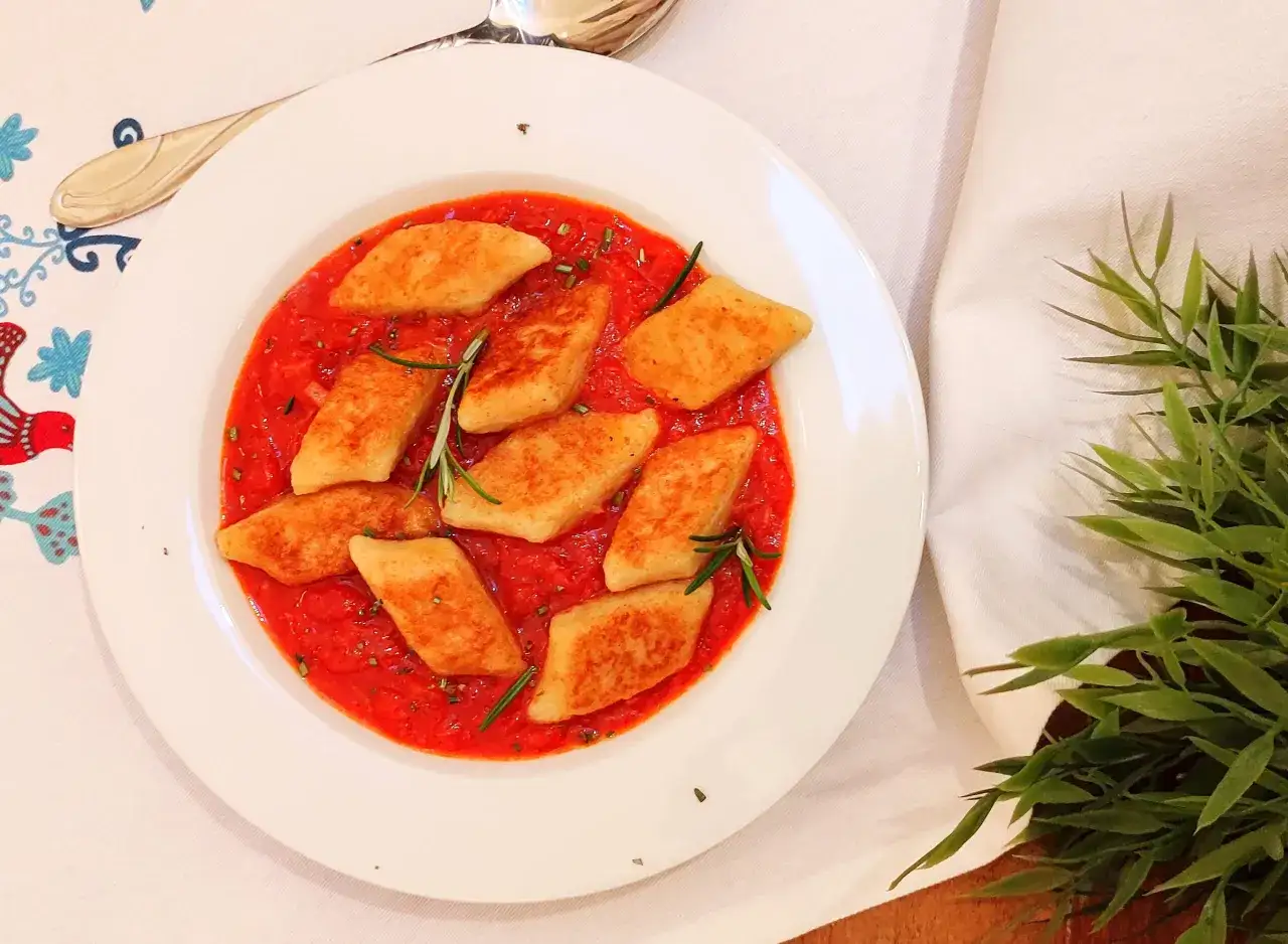 Kopytka w sosie pomidorowym z ziołami, podane na białym talerzu. Pyszny sos do kopytek zachęca do spr&oacute;bowania.