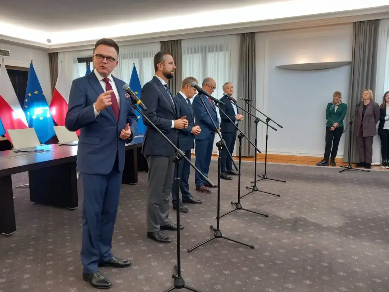 Mężczyźni w garniturach przemawiają przy mikrofonach. Na stole leży dokument z napisem 