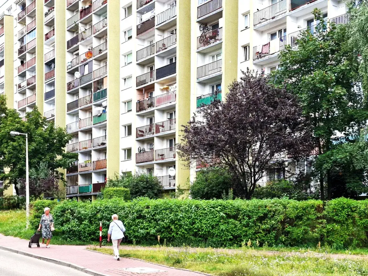 Wielka płyta z balkonami i zielenią. Widać fragmenty wentylacji w bloku z wielkiej płyty, a także spacerujące kobiety.