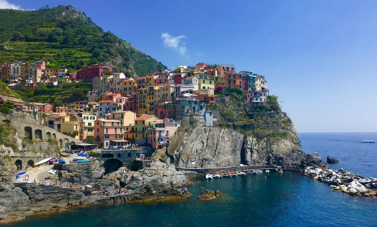 Kolorowe domy Cinque Terre wspinają się po klifie nad turkusowym morzem. W oddali widać łodzie, a na skałach odpoczywają ludzie.