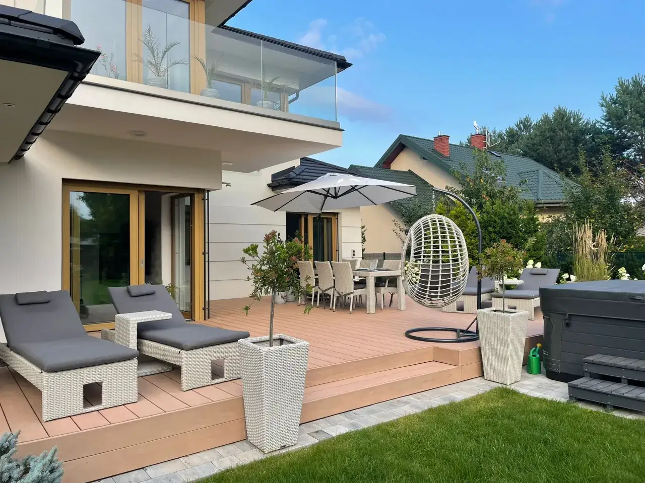 Nowoczesny taras przed domem z leżakami, stołem i krzesłami, wiszącym fotelem i jacuzzi. Idealne miejsce na relaks.