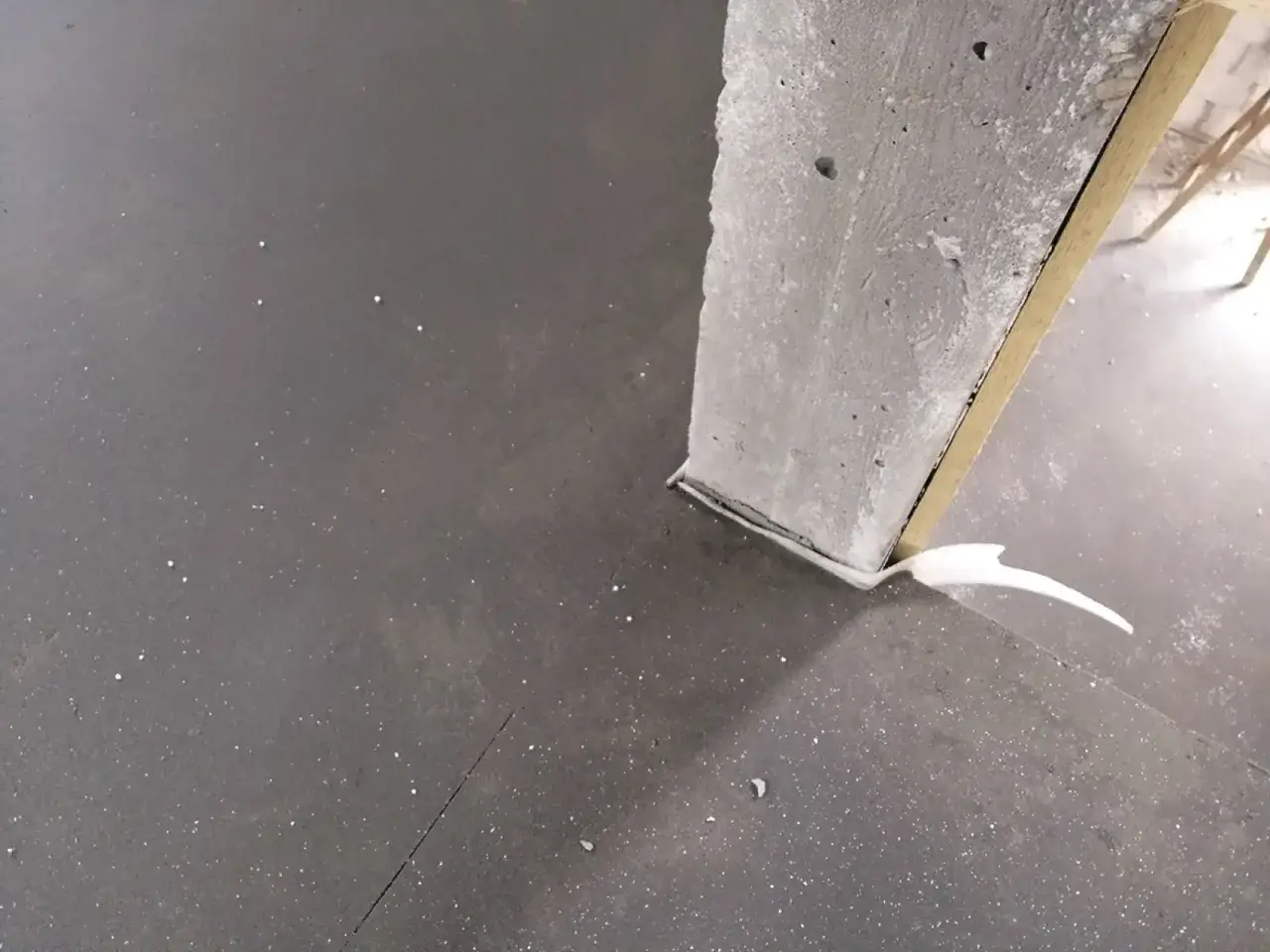 Szara, gładka podłoga z widocznymi drobnymi białymi kropkami. Obok betonowego słupa, który jest częścią budowy, widać białą piankę montażową. To etap prac co po wylewkach.
