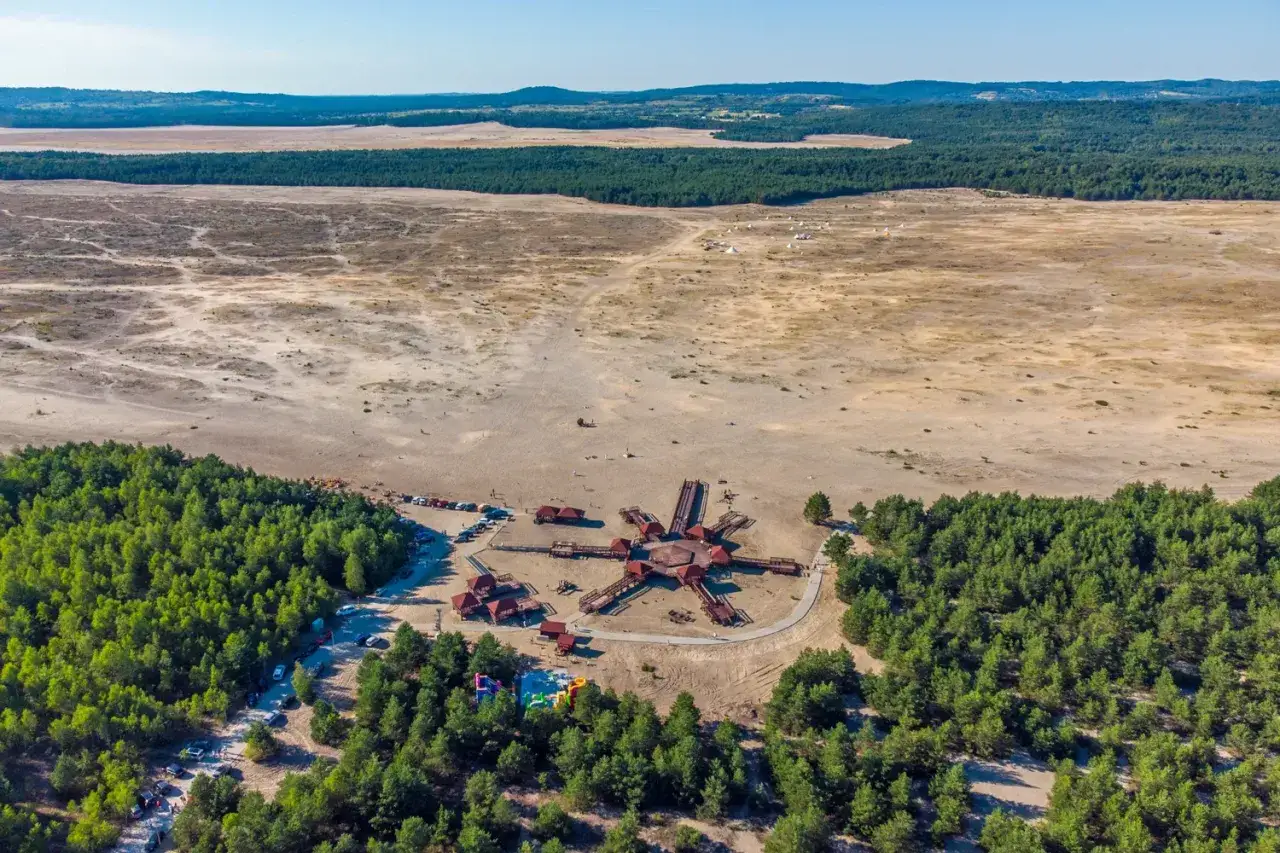 Pustynia Błędowska z lotu ptaka. Widoczna drewniana konstrukcja przypominająca gwiazdę, otoczona lasem. W oddali piaszczyste tereny i zielone wzgórza. Idealne miejsce na pustynia błędowska trasa rowerowa.