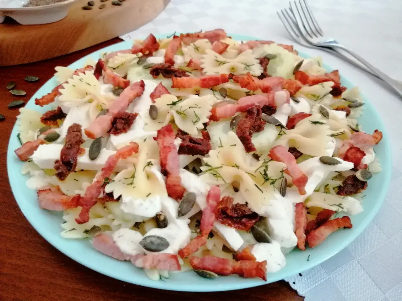 Pyszna sałatka z makaronem farfalle, chrupiącym boczkiem, serem i suszonymi pomidorami, posypana pestkami dyni.