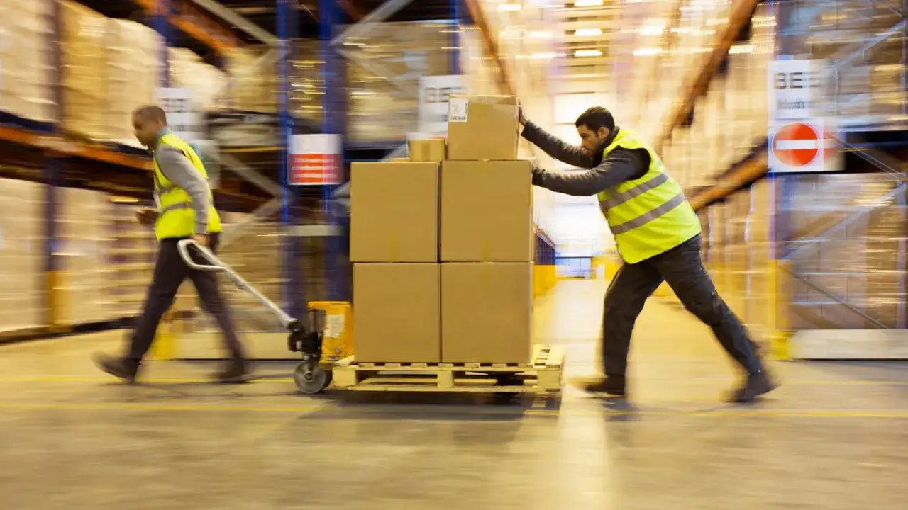 Dw&oacute;ch pracownik&oacute;w magazynu w kamizelkach odblaskowych. Jeden ciągnie w&oacute;zek z paczkami, drugi je pcha. Ruch i dynamika pracy.