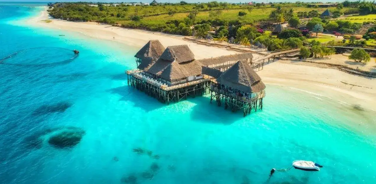 Rajskie Zanzibar ceny: domki na wodzie z krytymi strzechą dachami, turkusowa woda, biała plaża i bujna zieleń.