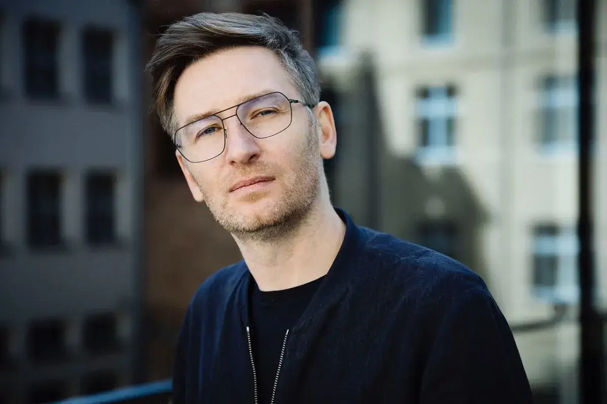 Portret Jarosława Tumidajskiego w okularach, z lekkim zarostem, na tle miejskiej zabudowy.