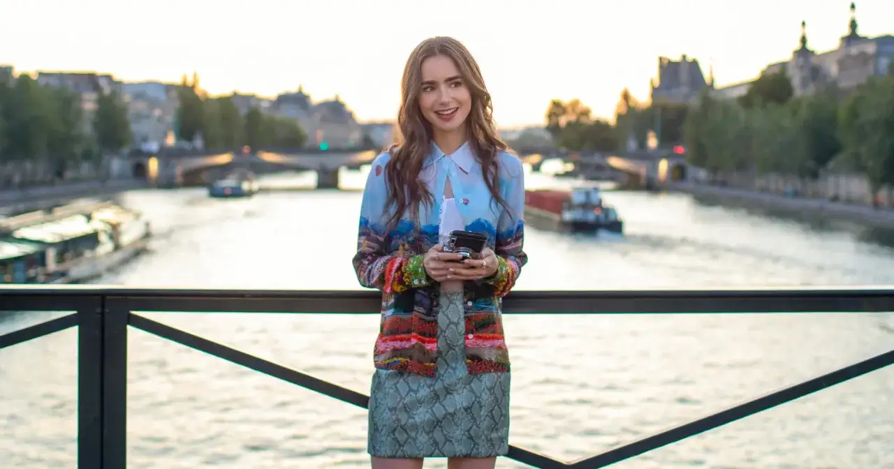 Emily w Paryżu z aparatem w ręku, na tle Sekwany i paryskich budynk&oacute;w.