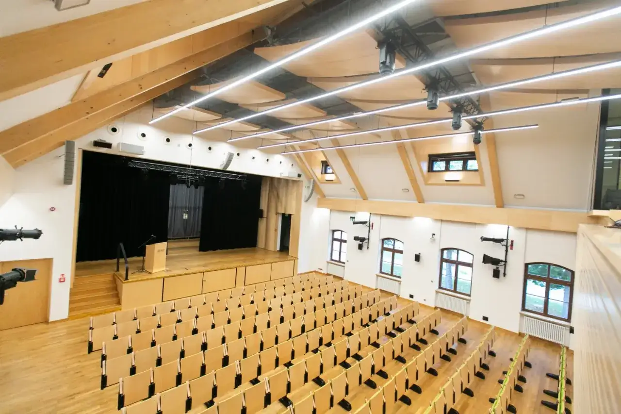 Nowa, nowoczesna aula z drewnianą sceną i rzędami krzeseł. Idealne miejsce na wykłady i wydarzenia.
