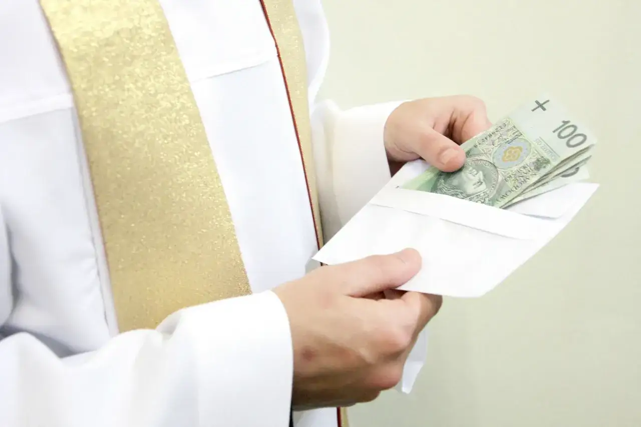 Dłoń w białej szacie z złotym pasem wkłada kopertę z banknotami. Pytanie: ile kosztuje chrzest w kościele?