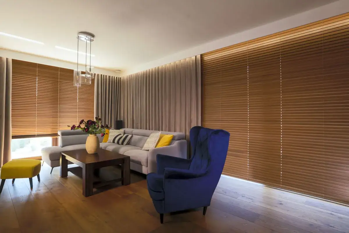 Elegancki salon z szarym narożnikiem, granatowym fotelem i ż&oacute;łtym pufem. W tle brązowe żaluzje drewniane i zasłony.