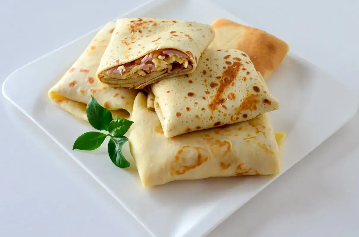 Złożone naleśniki z szynką i serem, jeden rozłożony, ukazujący pyszne nadzienie. Ułożone na białym talerzu z zielonym listkiem.