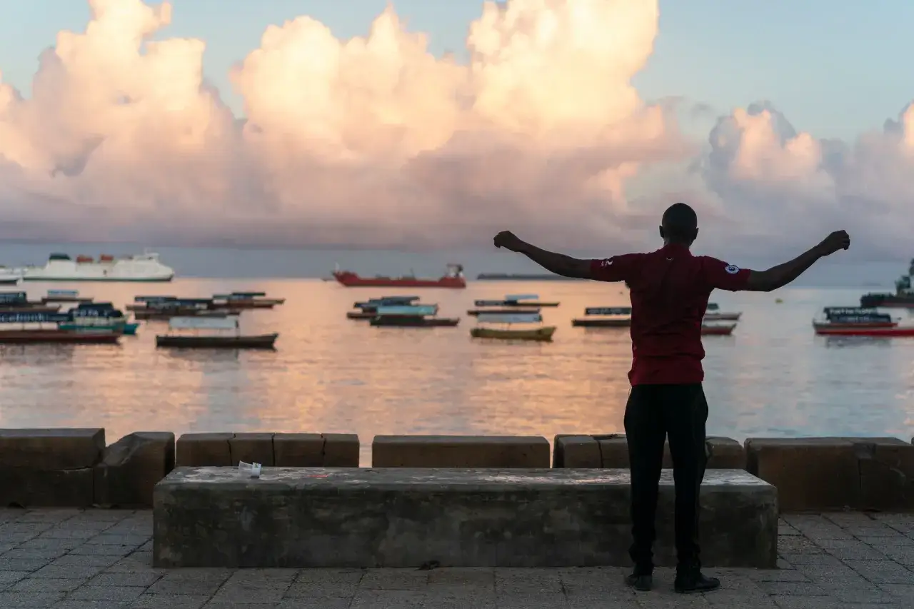 Mężczyzna z rozłożonymi ramionami wita dzień na Zanzibarze. W tle statki i łodzie na spokojnym morzu, chmury na niebie. To piękny widok, nawet jeśli zanzibar pora deszczowa przyniesie deszcz.