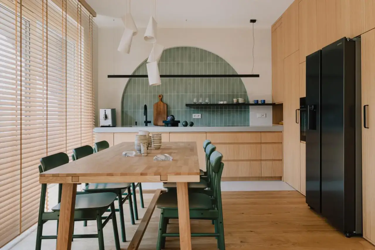Nowoczesna kuchnia z drewnianymi frontami i zielonymi płytkami tworzącymi łuk nad oknem. Drewniany st&oacute;ł z zielonymi krzesłami.