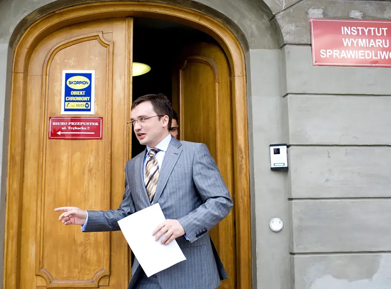 Jerzy Ziobro, w garniturze, wychodzi z budynku. Na drzwiach widnieje tabliczka 