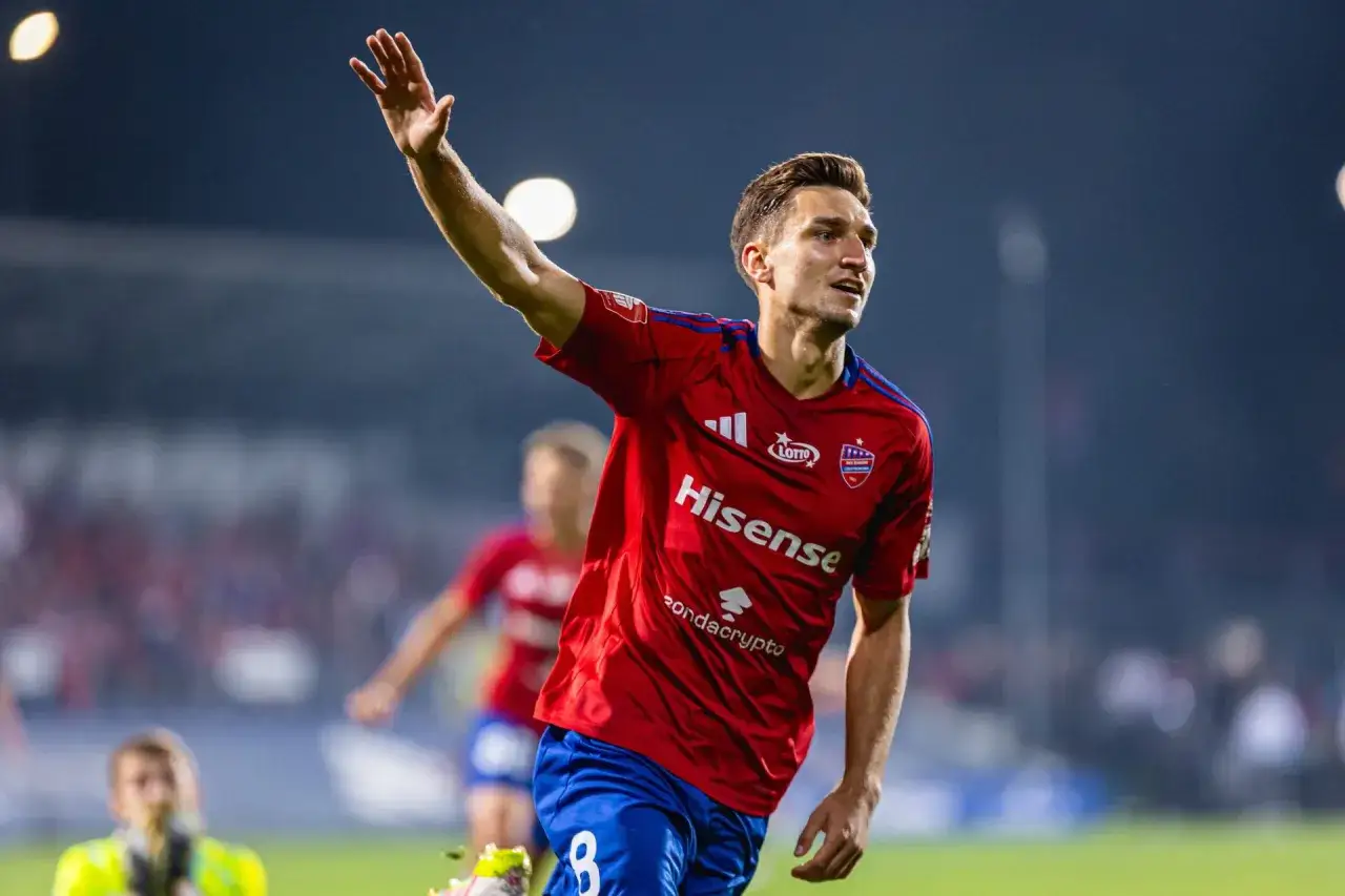 Piłkarz Rakowa Częstochowa w czerwonej koszulce z numerem 8, z uniesioną ręką, pozdrawia kibiców.