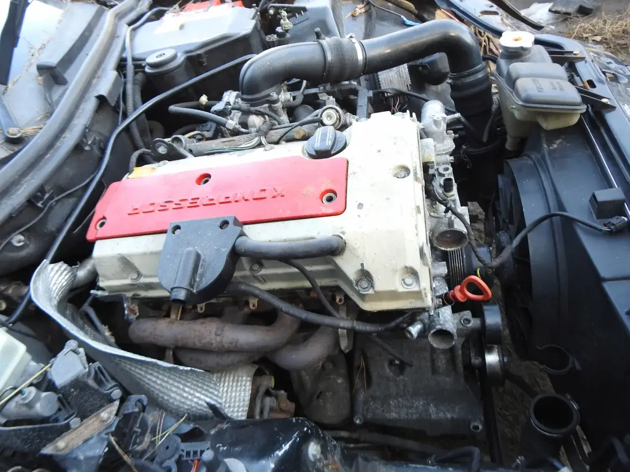 Silnik Mercedesa CLK W209 z czerwoną pokrywą kompresora. Widoczne przewody, wentylator i elementy układu chłodzenia.