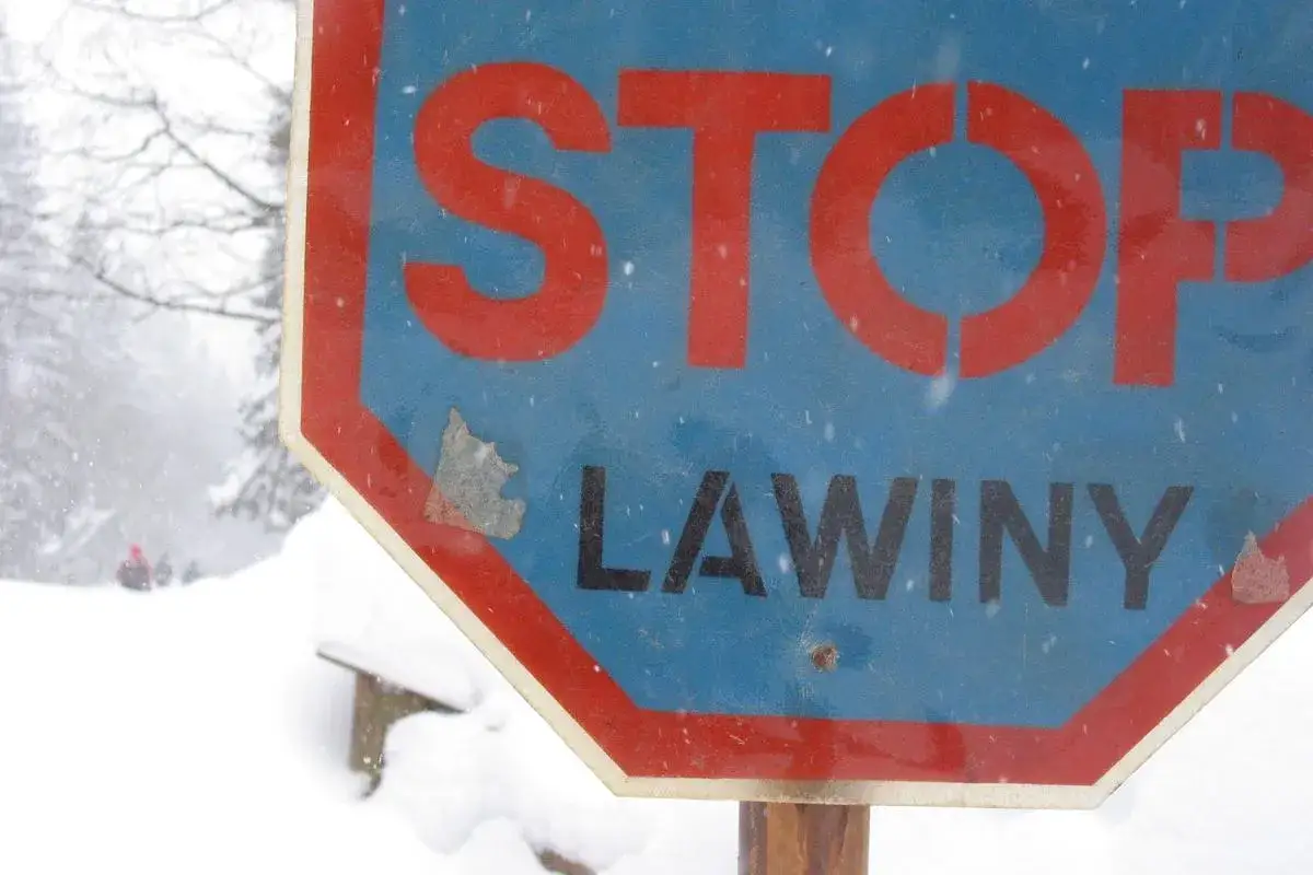 Znak STOP informuje o zagrożeniu lawinowym. W tle widać zaśnieżony krajobraz i ludzi w oddali.