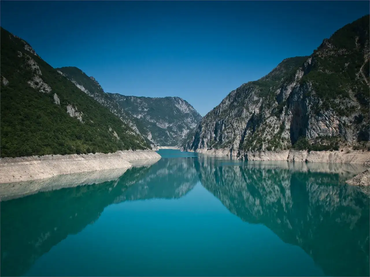 Turkusowe wody Pivsko Jezero odbijają skaliste g&oacute;ry porośnięte zielenią.