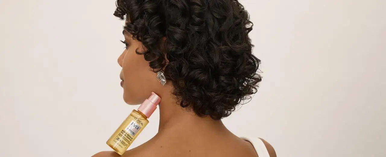 Kobieta z kręconymi włosami trzyma serum L'Oréal, które pomaga zwalczać rozdwojone końcówki i chroni włosy.
