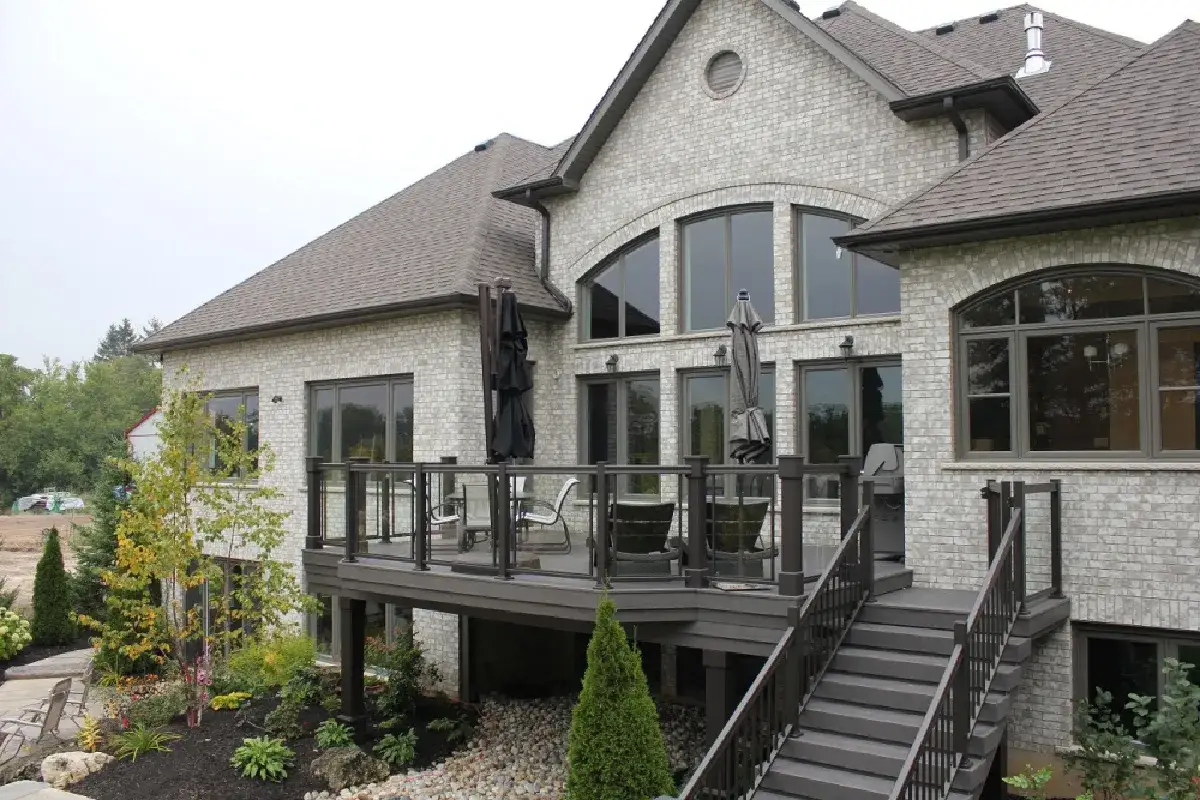 Wysoki taras na słupach z meblami i parasolami, prowadzący do domu z szarej cegły.