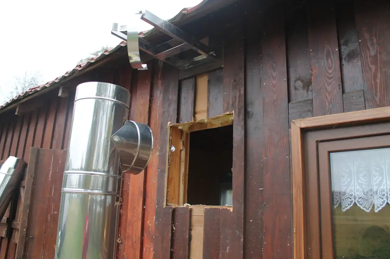 Metalowy komin przechodzi przez ścianę drewnianą. Widać otwór w ścianie i fragment okna z koronkową firanką.