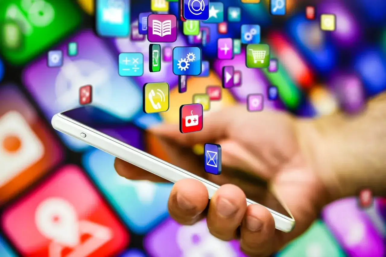 Dłoń trzyma smartfon, z którego wylatują kolorowe ikony aplikacji. W tle widać rozmyte, wielobarwne tło z innymi ikonami, sugerujące mnogość **aplikacji w tle**.