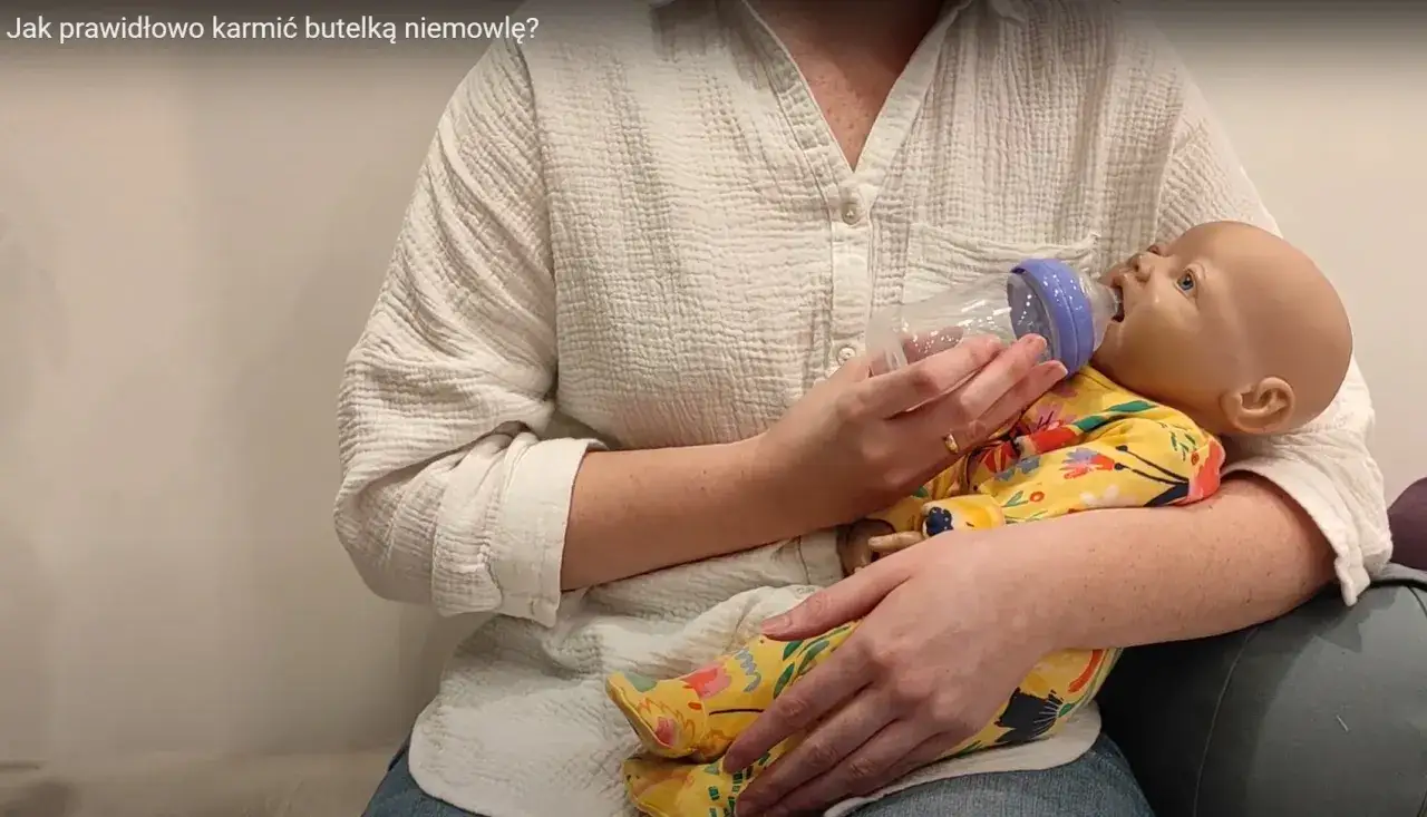 Osoba trzyma lalkę w odpowiedniej pozycji do karmienia butelką.