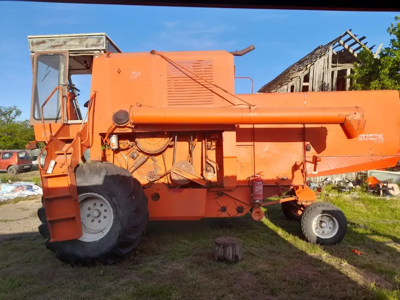 Pomarańczowy kombajn Bizon Z056. Zastanawiasz się, jaki olej hydrauliczny do bizona? Maszyna stoi na trawie, w tle ruiny stodoły.