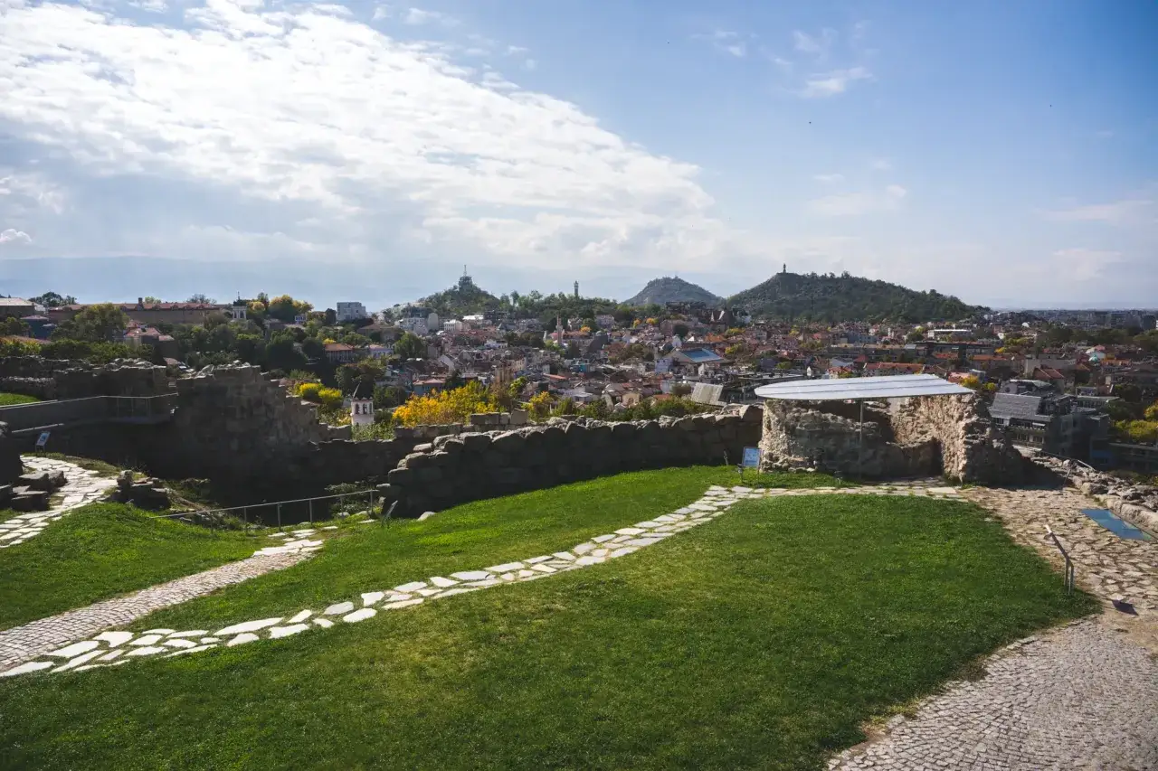 Widok na starożytne ruiny i miasto Plovdiv w Bułgarii. Kamienne ścieżki wiją się przez zielone trawniki, prowadząc do historycznych budowli.