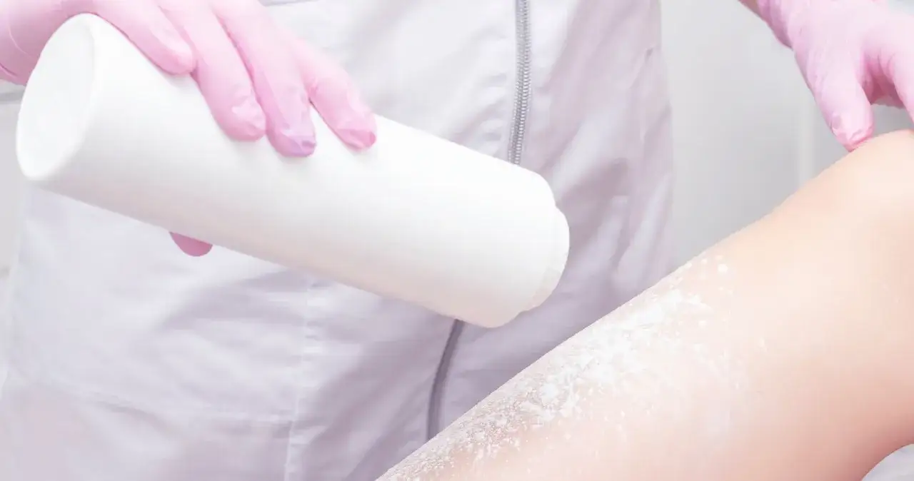 Ręce w r&oacute;żowych rękawiczkach sypią talk na nogę. To talk co to, przygotowanie do depilacji.