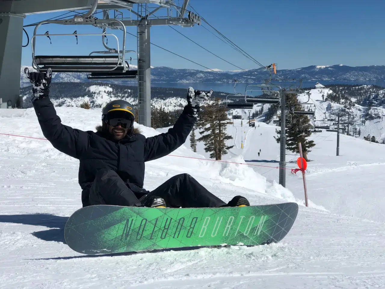 Uśmiechnięty snowboardzista na stoku, z rękami w górze. To zdjęcie przypomina o początkach snowboardingu, kiedy powstał ten sport.