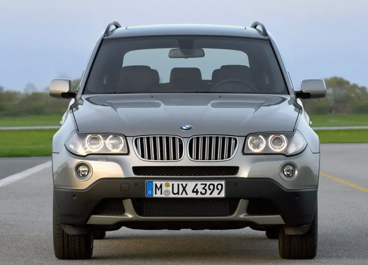 Szare BMW X3 E83 z włączonymi światłami, zaparkowane na zewnątrz.