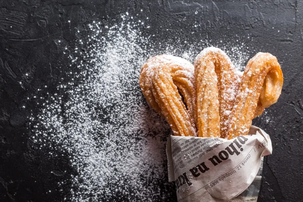 Churros z czekoladą kontra pączki z lukrem? Te złociste churros, posypane cukrem pudrem, kuszą na czarnym tle.