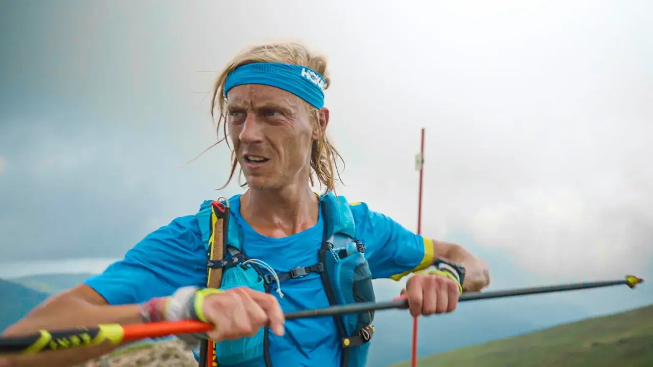 Roman Ficek, zmęczony, ale zdeterminowany biegacz g&oacute;rski, z niebieską opaską na głowie i plecakiem, trzyma kijki trekkingowe.