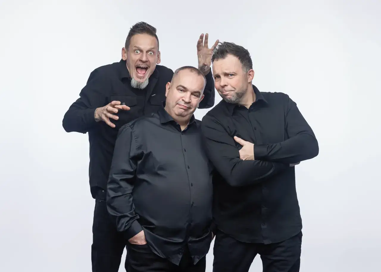 Trzej członkowie kabaretu Ani Mru Mru w czarnych koszulach pozują do zdjęcia. Jeden z nich robi śmieszną minę, drugi ma ręce skrzyżowane na piersi, a trzeci wyciąga ręce.