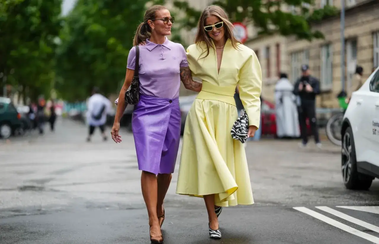 Dwie kobiety prezentują stylowe stylizacje ze sp&oacute;dnicą: jedna w fioletowej, błyszczącej, druga w ż&oacute;łtej, rozkloszowanej.