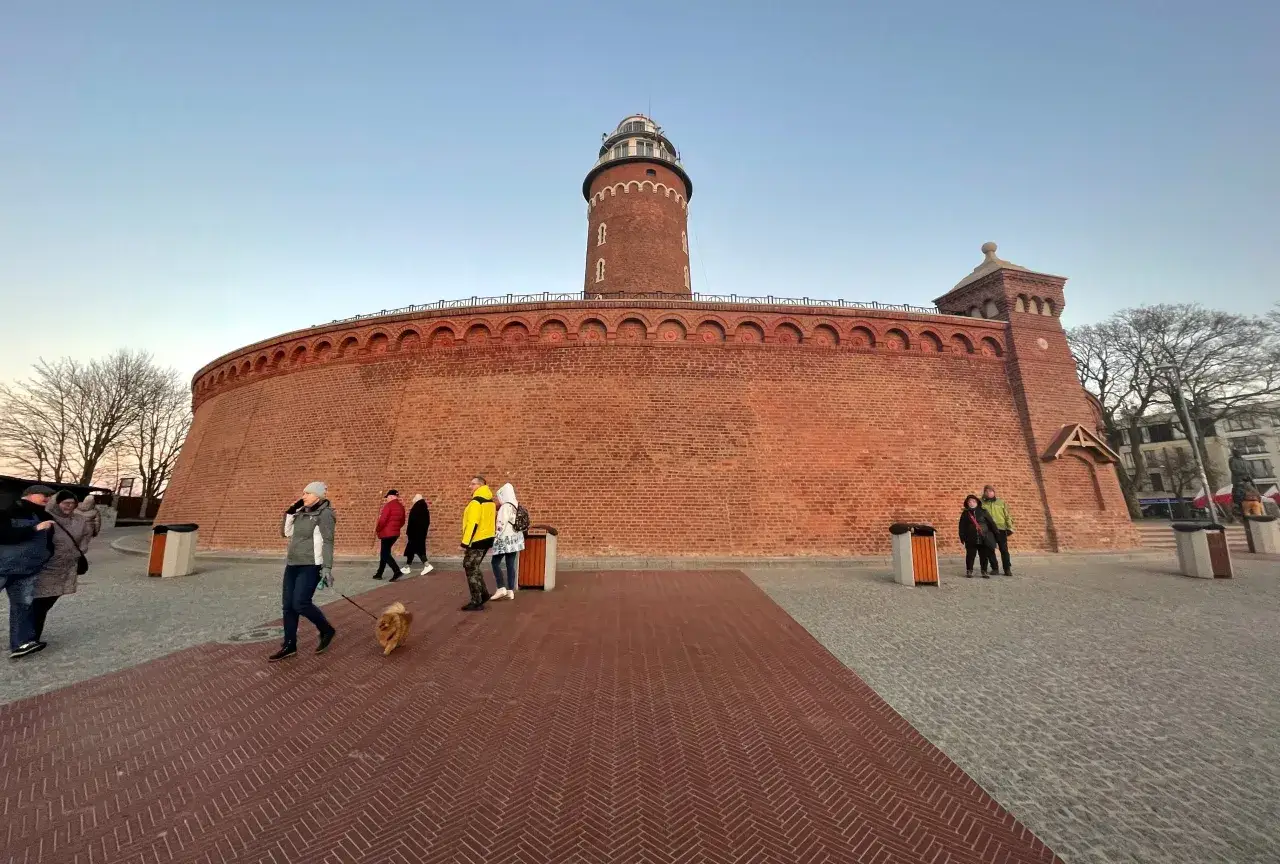 Latarnia morska w Kołobrzegu, imponująca ceglana budowla, to jedno z miejsc, co warto zobaczyć w Kołobrzegu. Spacerujący ludzie i pies dodają uroku.