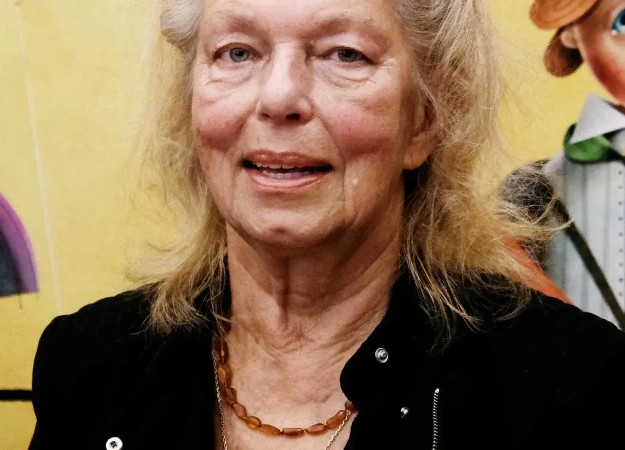 Uśmiechnięta Katarzyna Gaertner, z siwymi włosami i bursztynowym naszyjnikiem, obok postaci z bajki, jakby opowiadała swoim dzieciom historię.