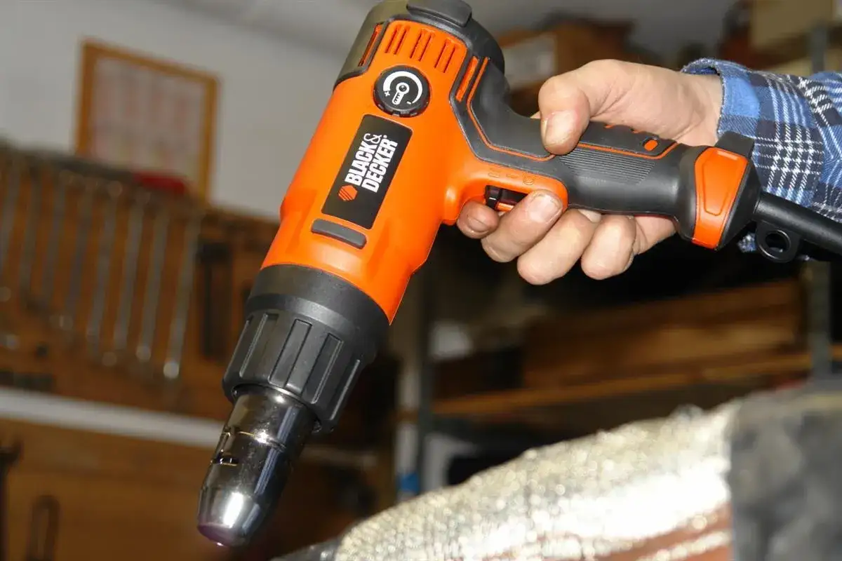 Pomarańczowa opalarka Black & Decker w dłoni, gotowa do pracy. W tle warsztat z narzędziami.