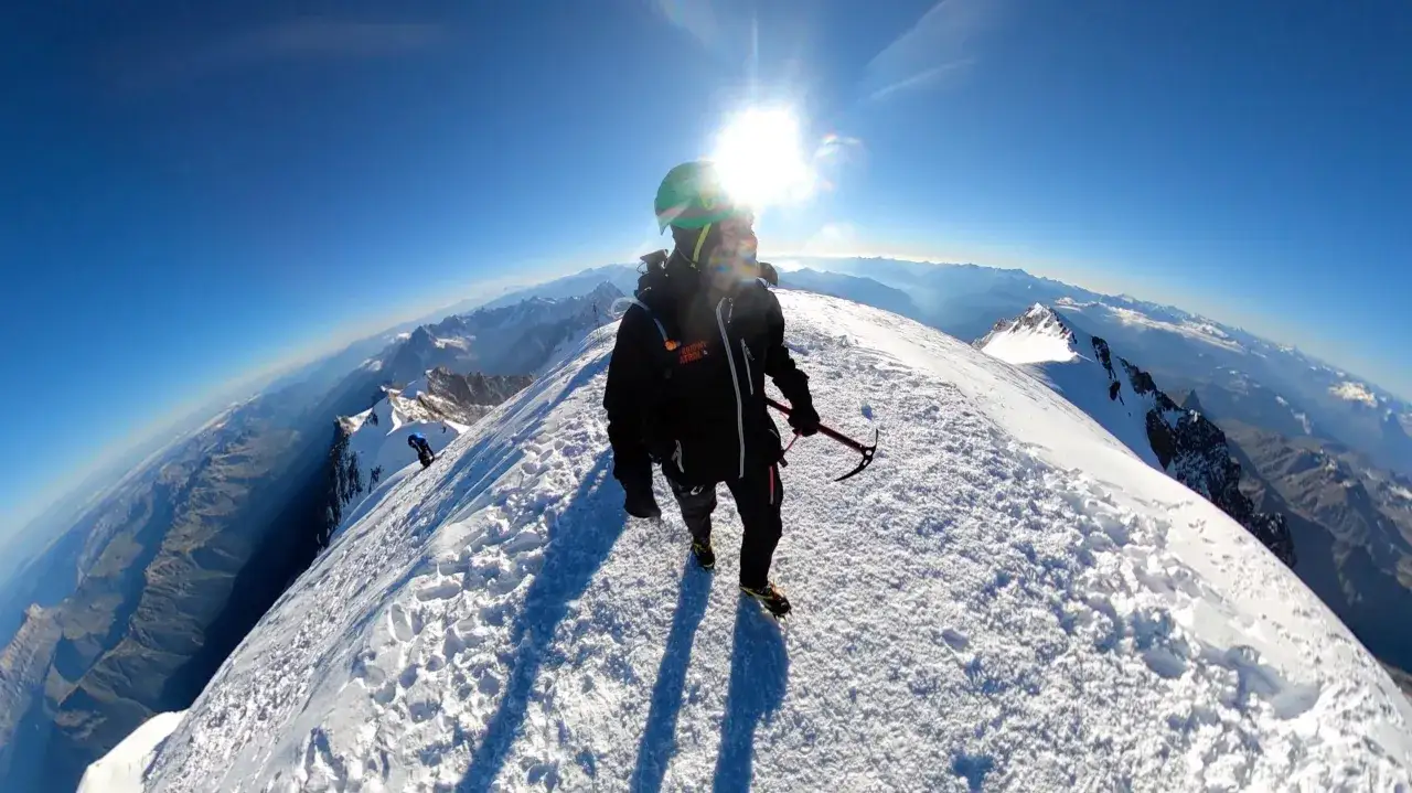 Alpinista na śnieżnym grzbiecie, z czekanem w dłoni, podziwia widok z najwyższego szczytu Alp. Słońce świeci jasno na błękitnym niebie.