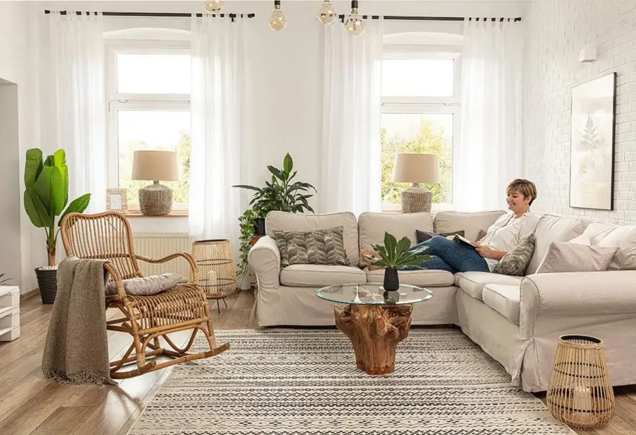 Przytulny, mały pokój w stylu boho z wiklinowym fotelem, jasną kanapą i dużą ilością zieleni. Kobieta czyta książkę.