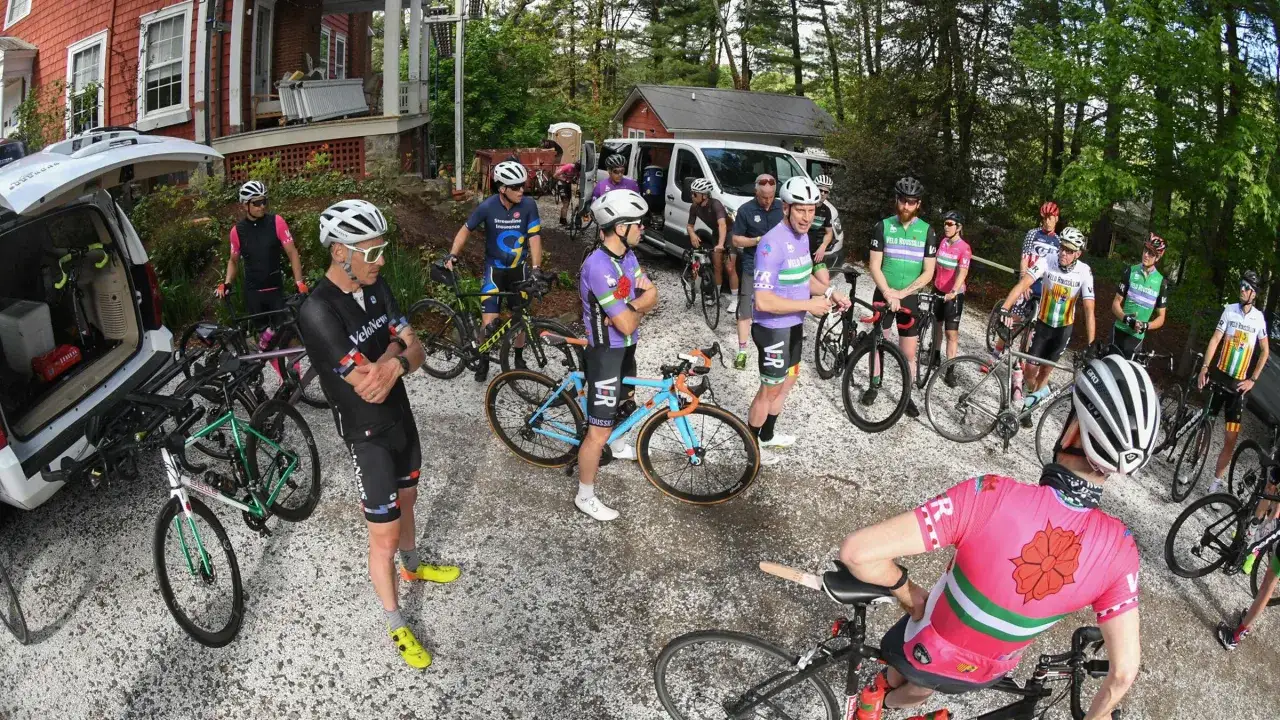 Grupa kolarzy szosowych w kolorowych strojach przygotowuje się do wyścigu. Rowery stoją obok samochod&oacute;w.