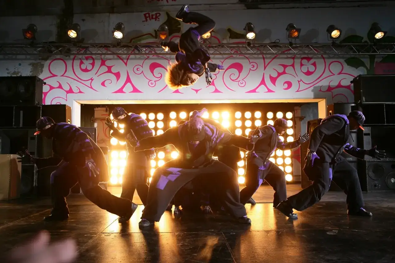 Tancerze w strojach z fioletowymi akcentami wykonują dynamiczne ruchy, jeden z nich akrobatycznie wiruje w powietrzu. Scena jak z film&oacute;w o tańcu ulicznym.