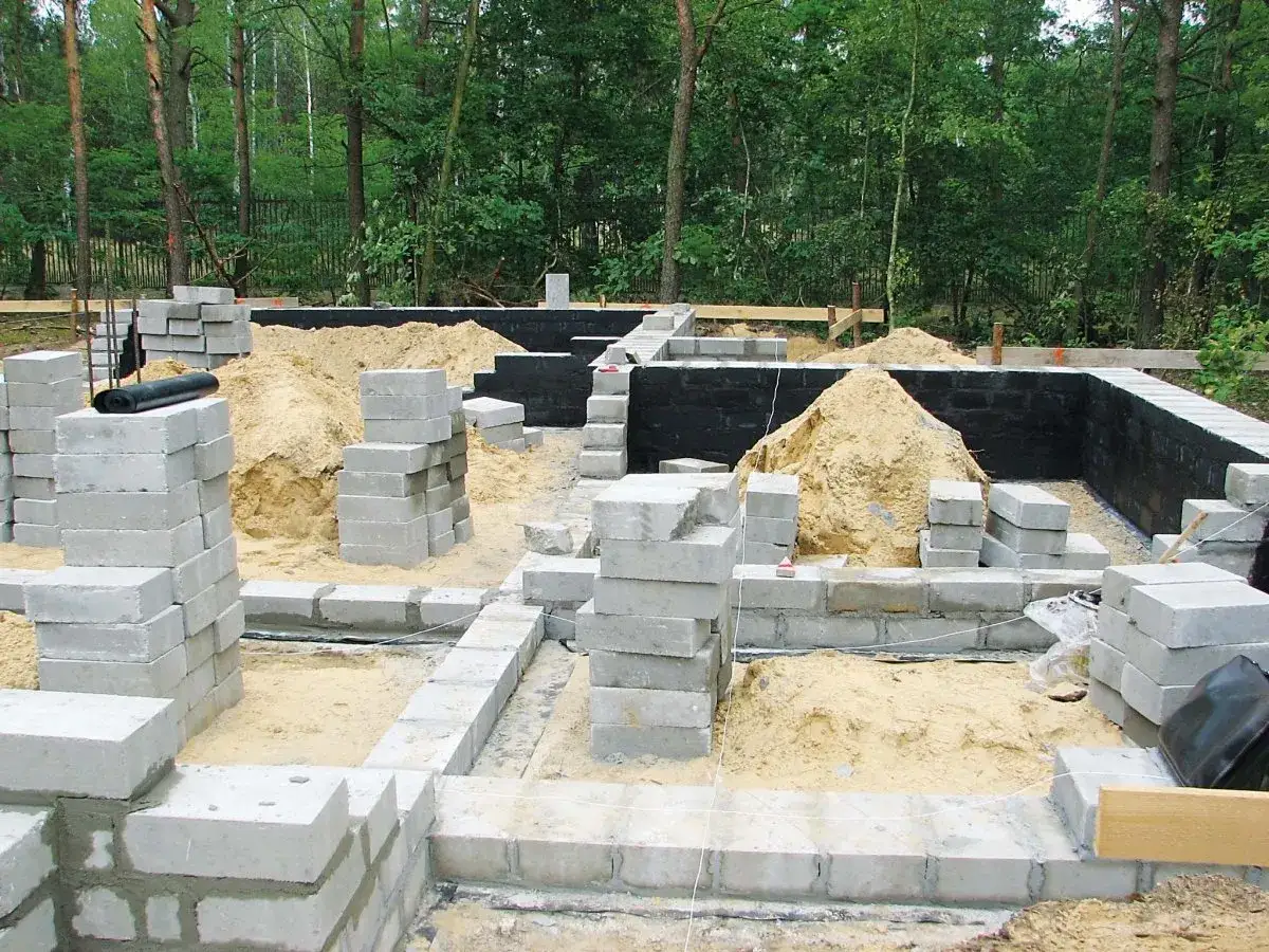 Budowa fundamentów z bloczków betonowych. W tle piasek i czarna izolacja. Zastanawiasz się, jaki cement na fundamenty będzie najlepszy?